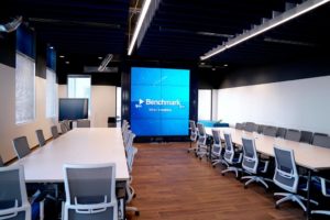 Large meeting room with two long tables (chairs surrounding both) and a large media screen at the back of the room